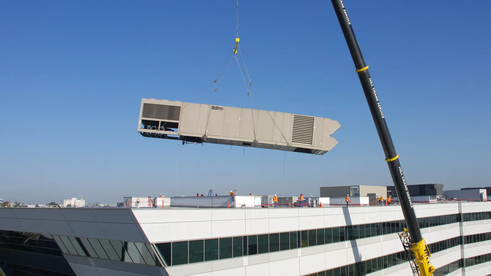 A packaged rooftop HVAC unit lifted by crane over an office building roof, the usual way rooftop equipment moves in a commercial HVAC replacement